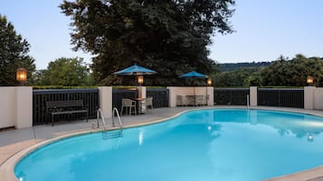 Ein Schwimmbecken mit einer umlaufenden Sonnenterrasse, zwei Sonnenschirmen und einem Tisch mit Stühlen.