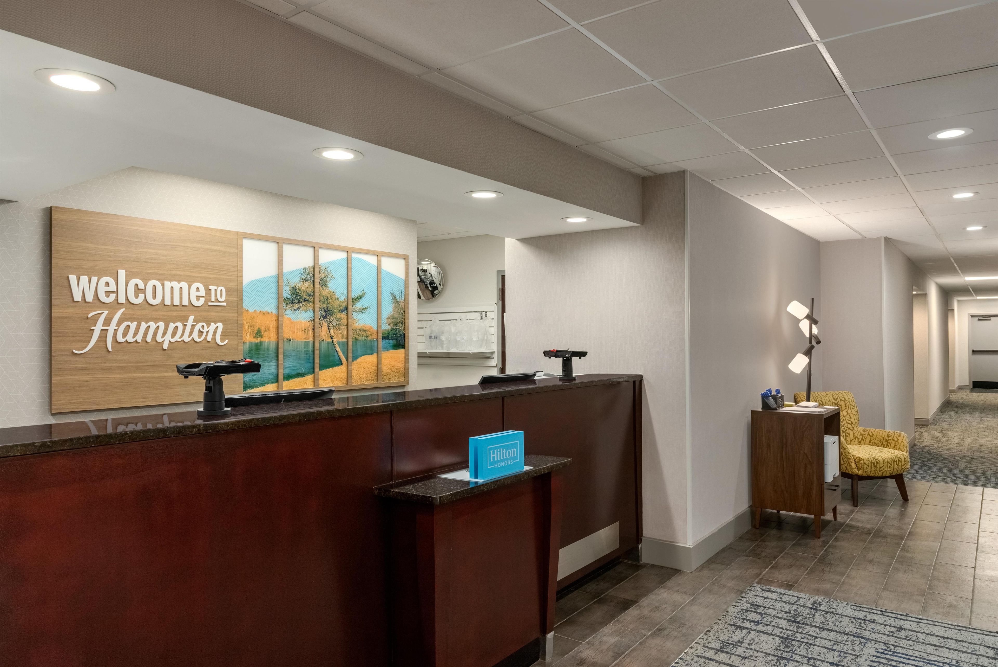 Ein Hotel-Empfangsbereich mit einem Holzbüro, einem Willkommensschild, einem gerahmten Landschaftsbild, einer Uhr und einer Sitzecke mit einer Lampe.