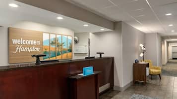 Ein Hotel-Empfangsbereich mit einem Holzbüro, einem Willkommensschild, einem gerahmten Landschaftsbild, einer Uhr und einer Sitzecke mit einer Lampe.