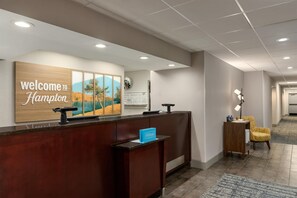 A hotel reception area with a wooden reception desk, a welcome sign, a framed landscape painting, a clock, and a seating area with a lamp. - Hampton Inn Bristol (Bristol)