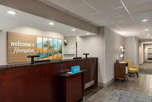 Un área de recepción de hotel con un mostrador de madera, un cartel de bienvenida, un cuadro de paisaje enmarcado, un reloj y un espacio de descanso con una lámpara.