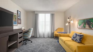 Habitación de hotel con un escritorio, un sofá amarillo, una silla, un televisor, un cuadro y una ventana con cortinas.