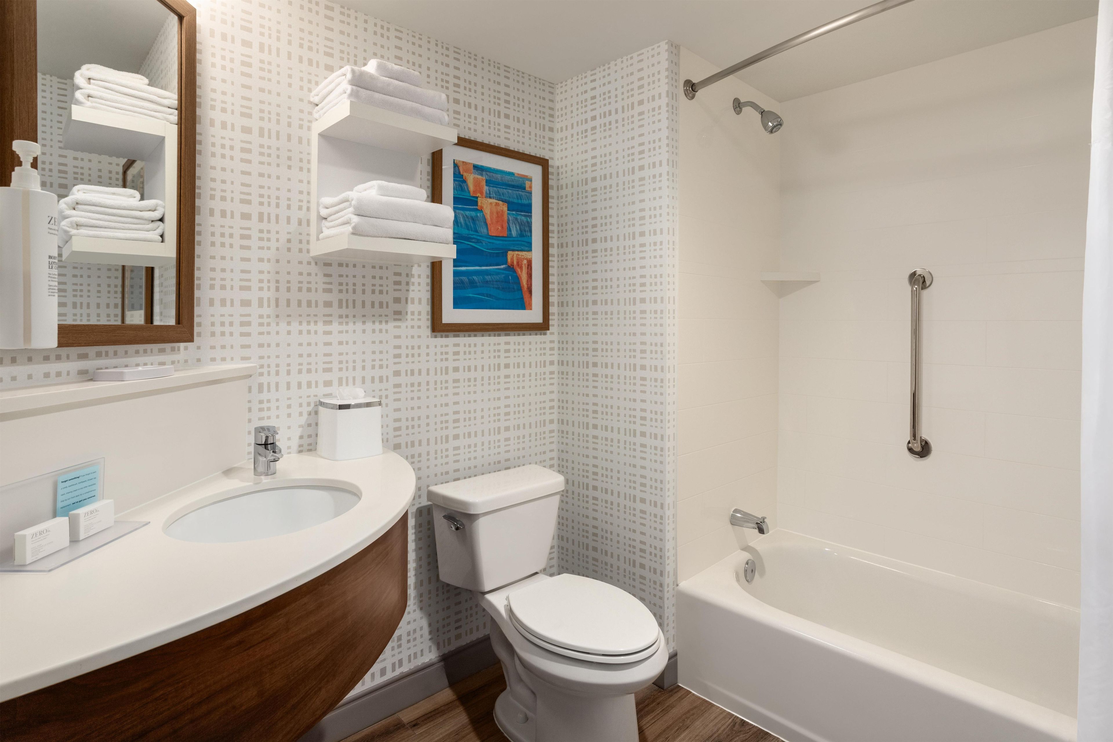 A bathroom with a sink, toilet, and bathtub. There are towels on a shelf and a framed picture on the wall.