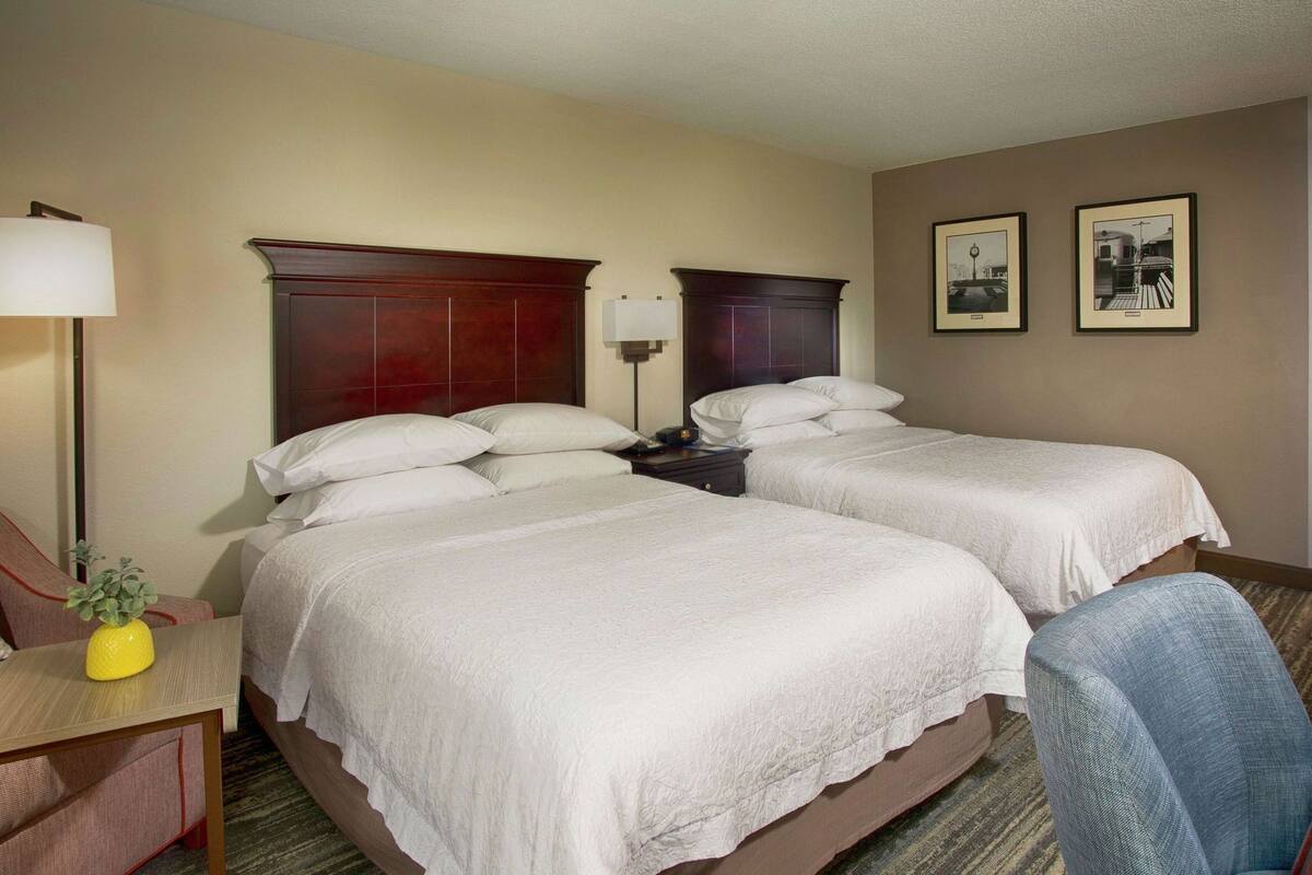 A hotel room with two beds, a desk with a lamp, a chair, and two framed pictures on the wall.