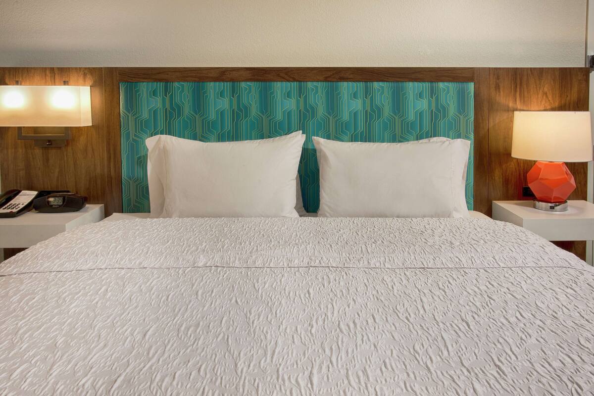 A hotel room with a large bed, two white pillows, a teal patterned headboard, a wooden accent, a red lamp, and a nightstand with a phone and a lamp.