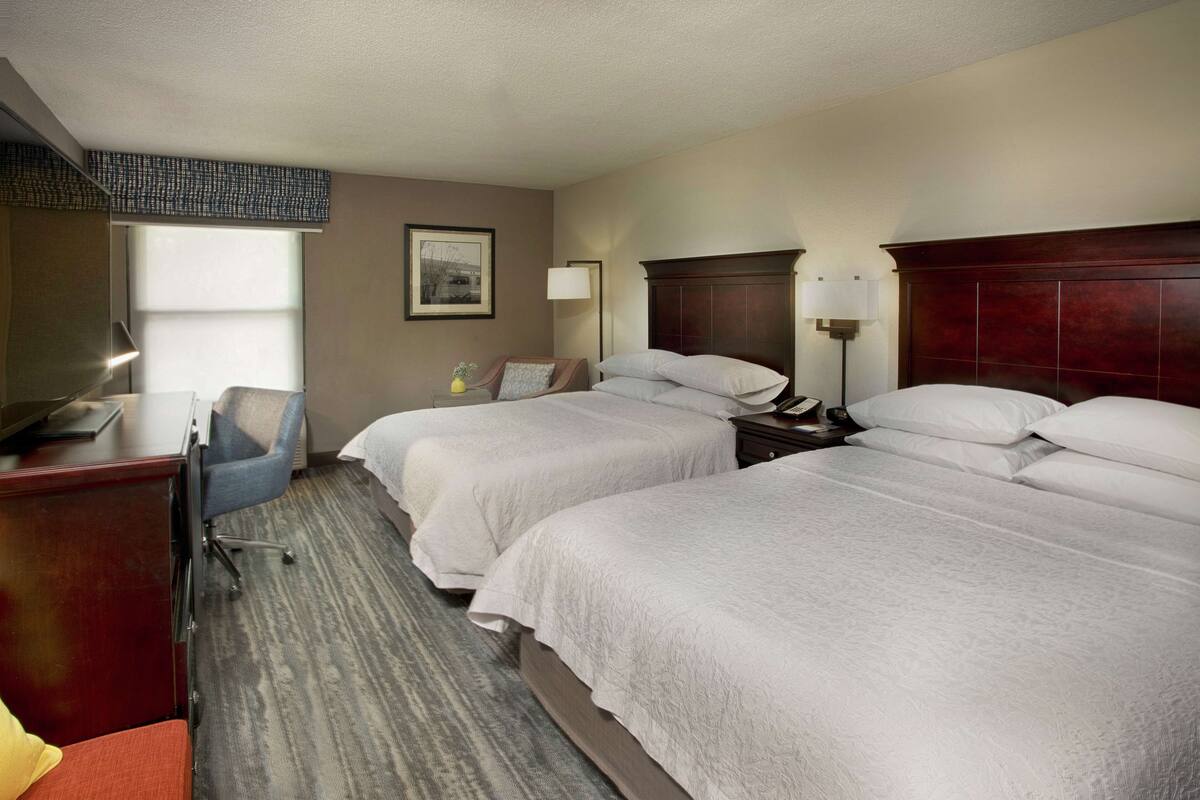 A hotel room with two beds, a desk, a chair, and a lamp.
