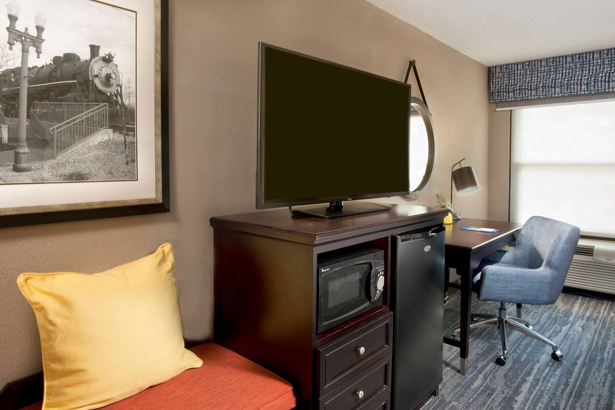 A hotel room with a bed, a desk with a chair, a television, a microwave, and a framed picture on the wall.