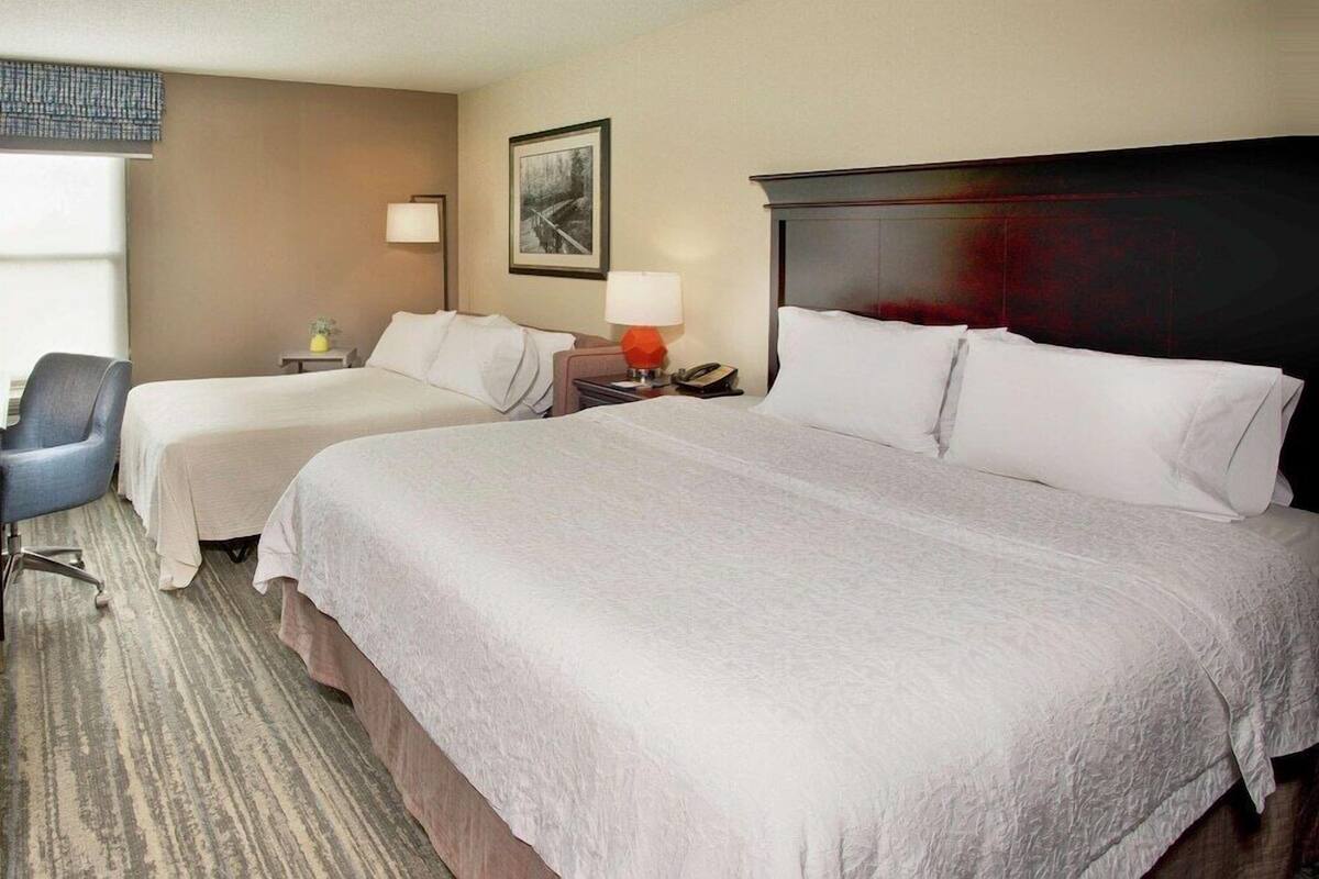 A hotel room with two beds, a desk, a chair, a lamp, and a framed picture on the wall.