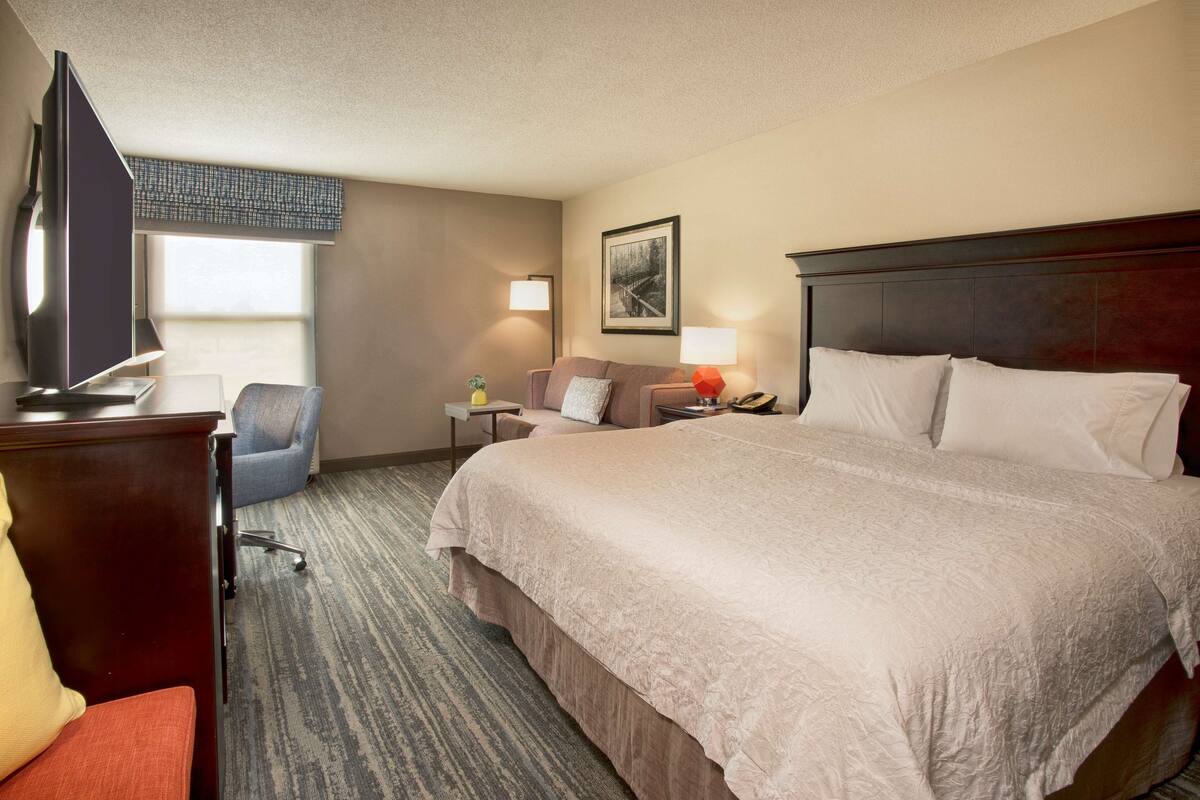 A hotel room with a large bed, a desk with a chair, a television, and a sofa.