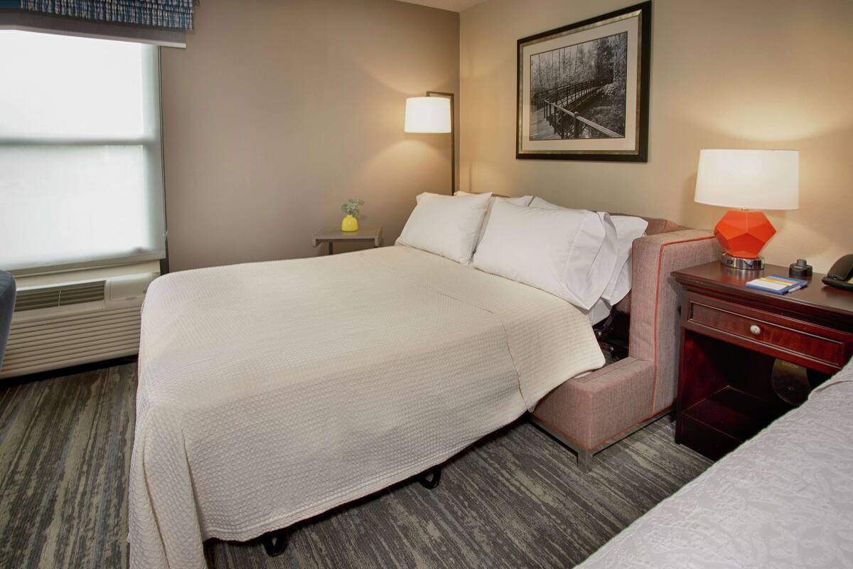 A hotel room with a large bed, a nightstand with a lamp, a small table with a vase, and a framed picture on the wall.
