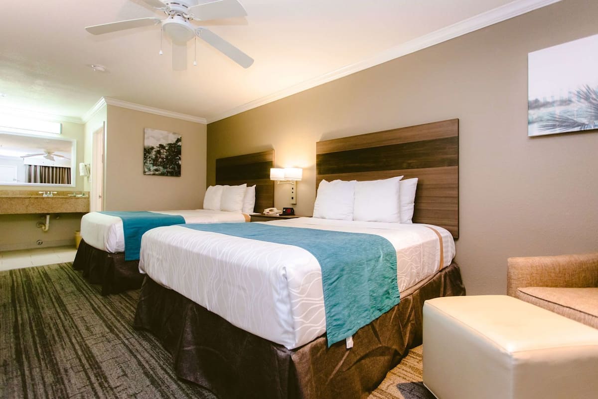 A hotel room with two beds, a ceiling fan, a nightstand with a lamp, a painting on the wall, and a view of the city.