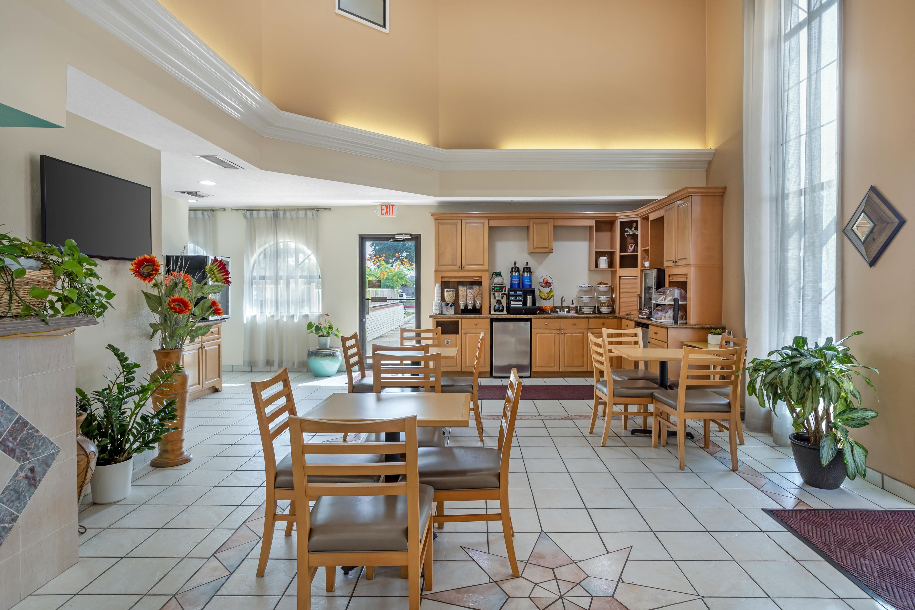 Uma área de refeições de hotel com uma mesa central, cadeiras e uma cozinha ao fundo.