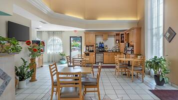Uma área de refeições de hotel com uma mesa central, cadeiras e uma cozinha ao fundo.