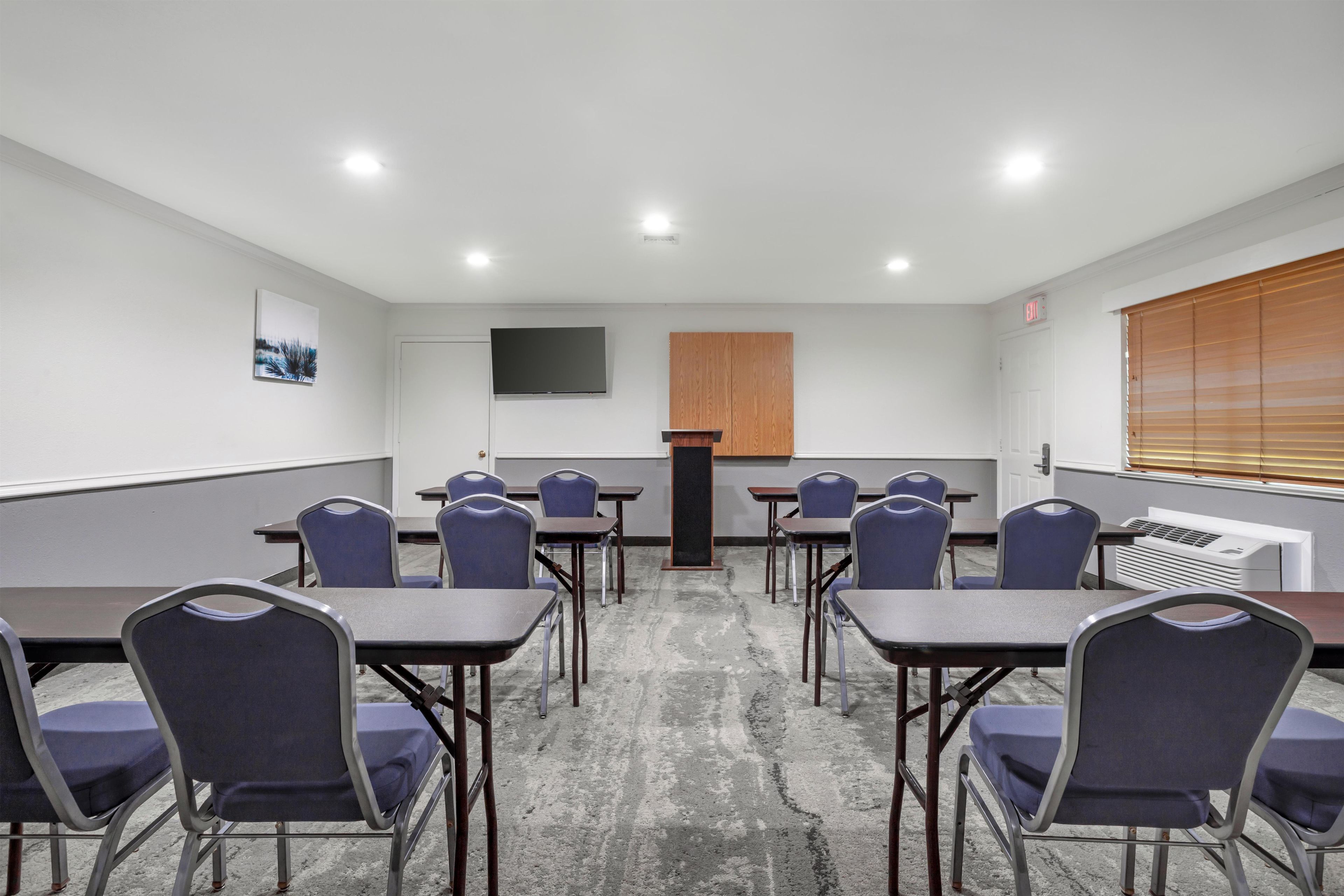 A conference room with tables and chairs, a flat-screen TV, a wooden panel, and a window with blinds.