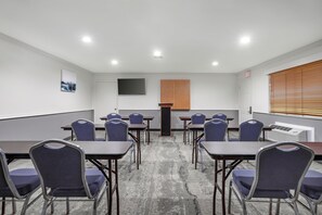 Un salón de conferencias con mesas y sillas, un televisor de pantalla plana, un panel de madera y una ventana con persianas.