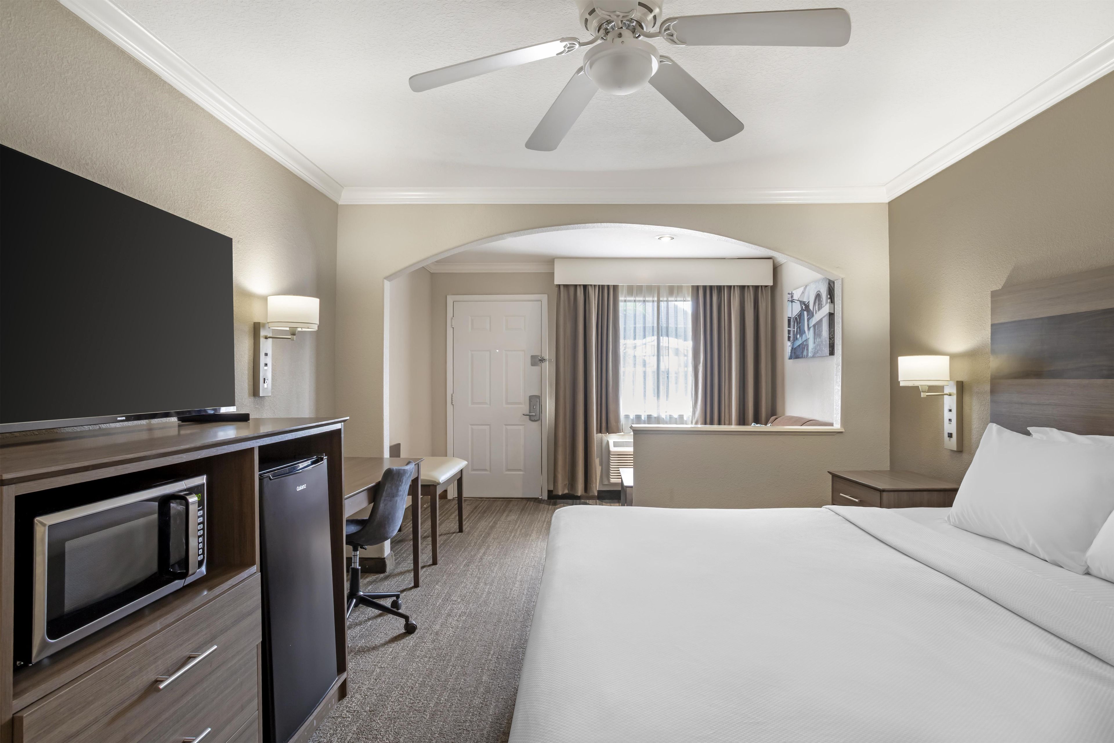 Quarto de hotel com cama, escrivaninha, televisão e ventoinha no teto.