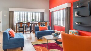 Una sala de estar moderna con comedor, que incluye un sofá azul, un sillón naranja y una mesa de centro con objetos decorativos.