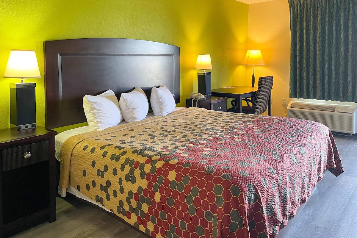 A hotel room with a large bed, two bedside tables with lamps, a desk with a chair, and an air conditioning unit.