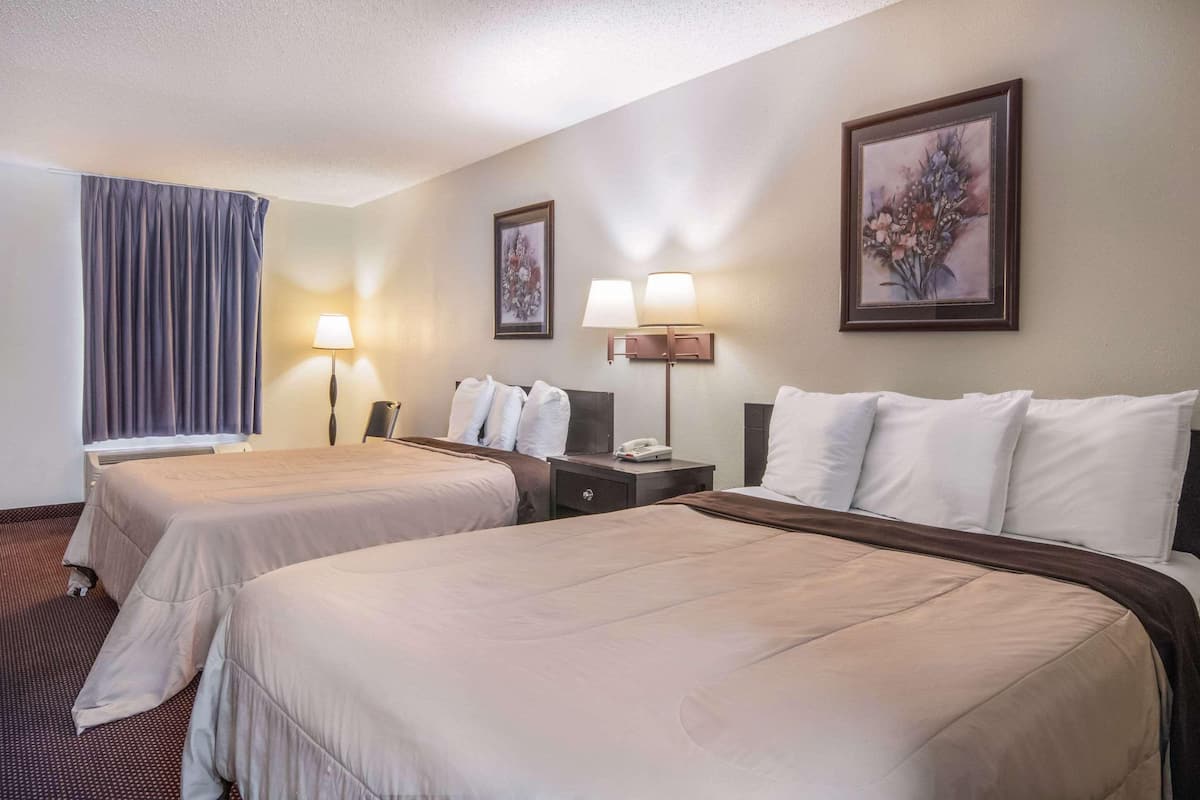 A hotel room with two beds, a nightstand with a lamp, a painting on the wall, and a window with curtains.