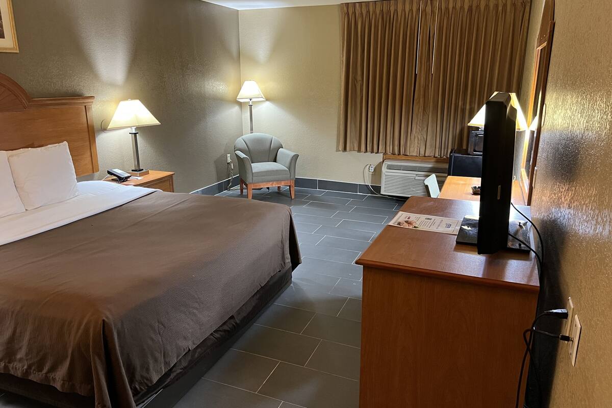 A hotel room with a large bed, a desk with a computer, a chair, two lamps, and a window with curtains.