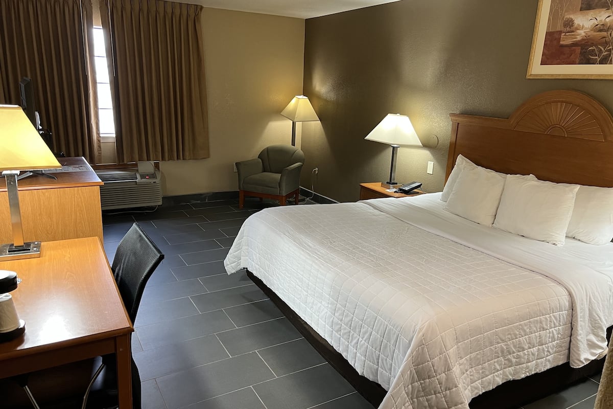 A hotel room with a bed, a desk, a chair, a lamp, and a painting on the wall.