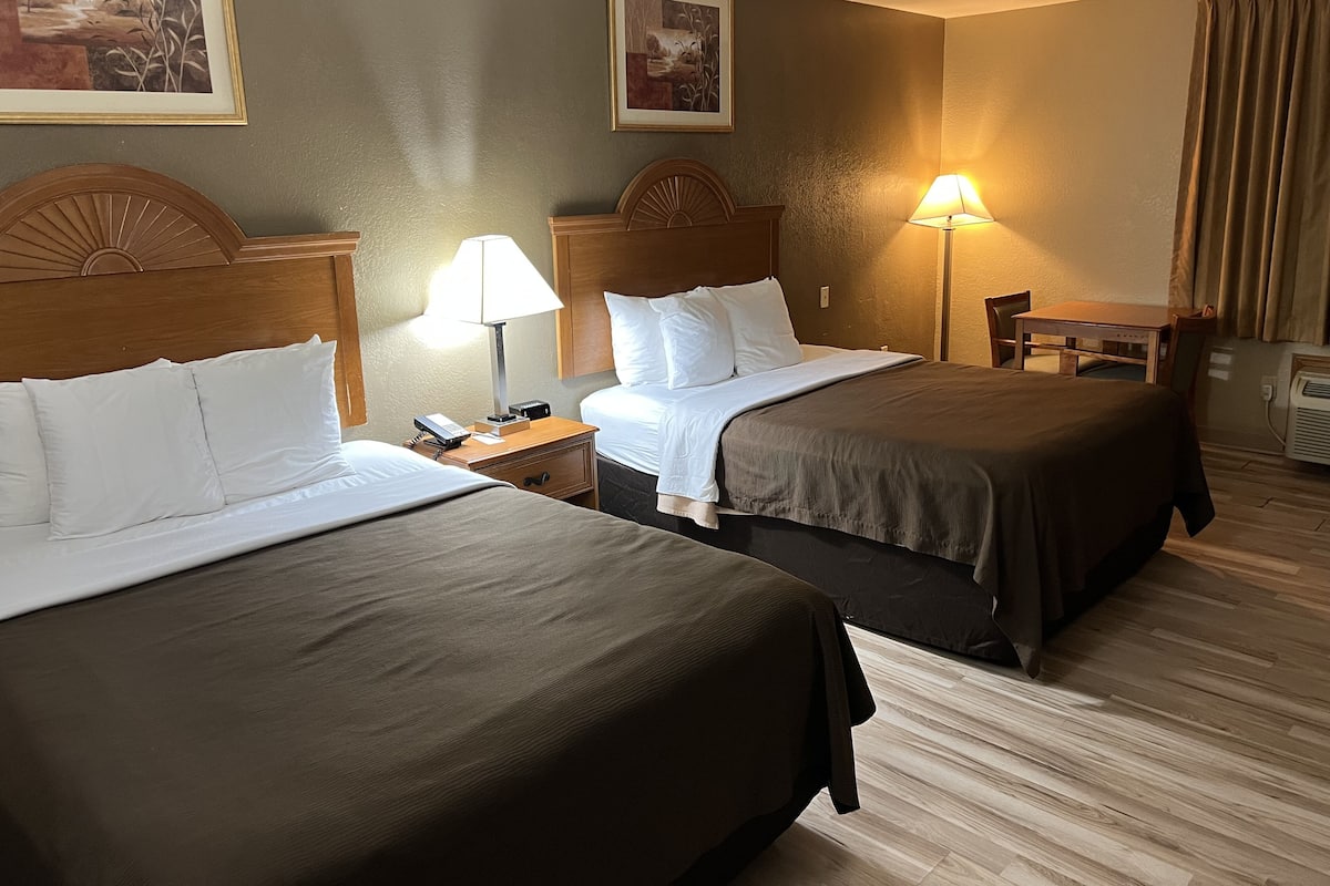 A hotel room with two beds, a wooden headboard, a nightstand with a lamp, and a desk with a chair.