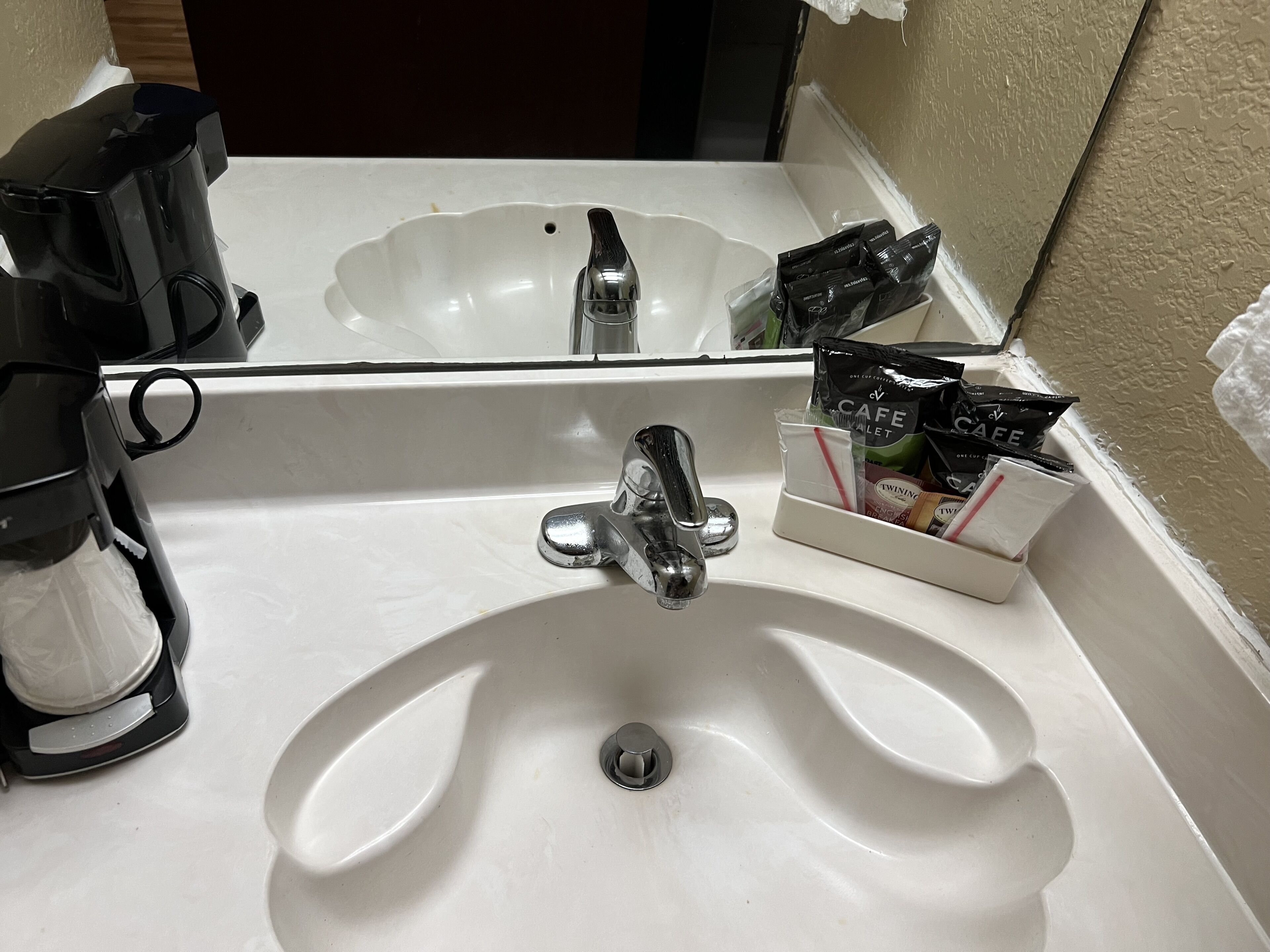 Un lavabo blanc dans la salle de bain, avec un miroir au-dessus, une machine à café à gauche et un distributeur d’articles de toilette à droite.