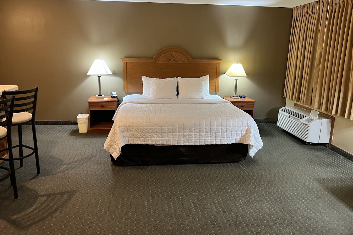 A hotel room with a large bed, two bedside tables with lamps, a chair, a waste bin, and a wall-mounted air conditioner.