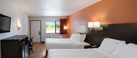 A hotel room with two beds, a desk, a TV, and a window with a view of trees.