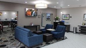A hotel lobby with blue sofas, a coffee station, and a TV screen displaying a news program. - Comfort Suites Lewisville - Dallas (Lewisville)