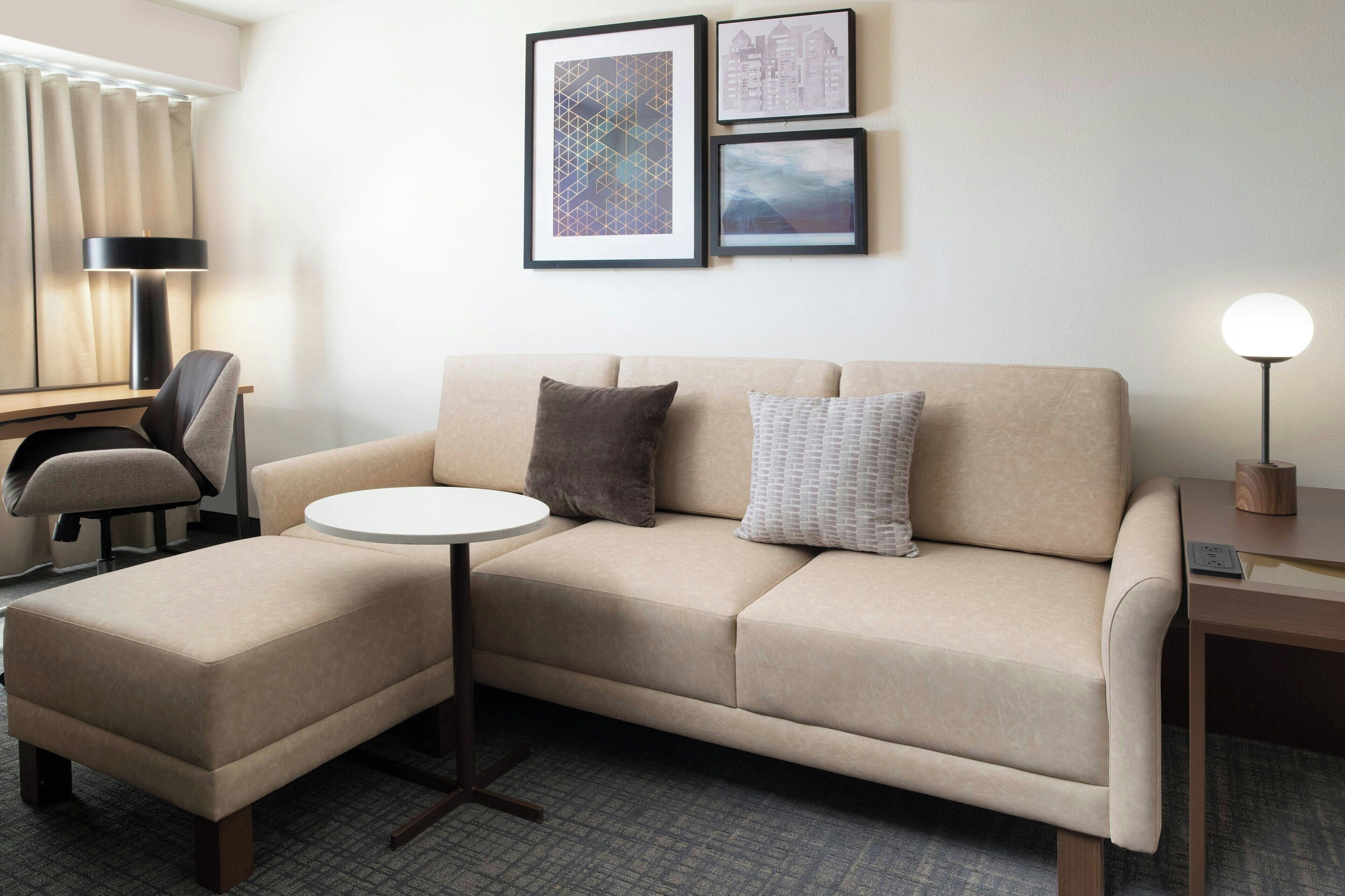 A modern hotel room with a beige sofa, a small round table, a desk with a chair, and framed artwork on the wall.