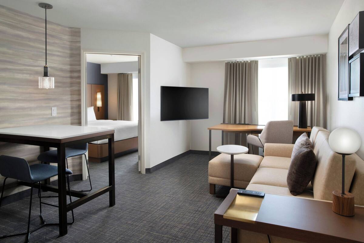 Habitación de hotel con televisor de pantalla plana, un sofá, una mesa de comedor con sillas, una mesita redonda y una lámpara.