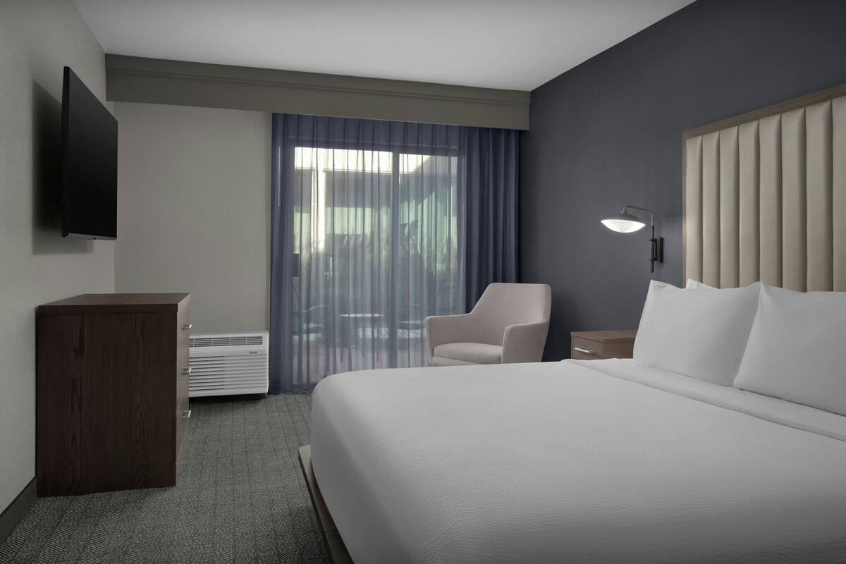 A hotel room with a large bed, a desk, a chair, a flat-screen TV mounted on the wall, and a window with curtains.