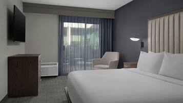 A hotel room with a large bed, a desk, a chair, a flat-screen TV mounted on the wall, and a window with curtains.