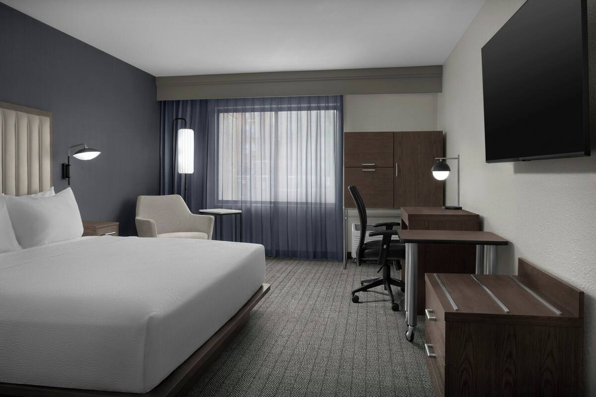 A hotel room with a large bed, a desk, a chair, a TV, and a window with curtains.