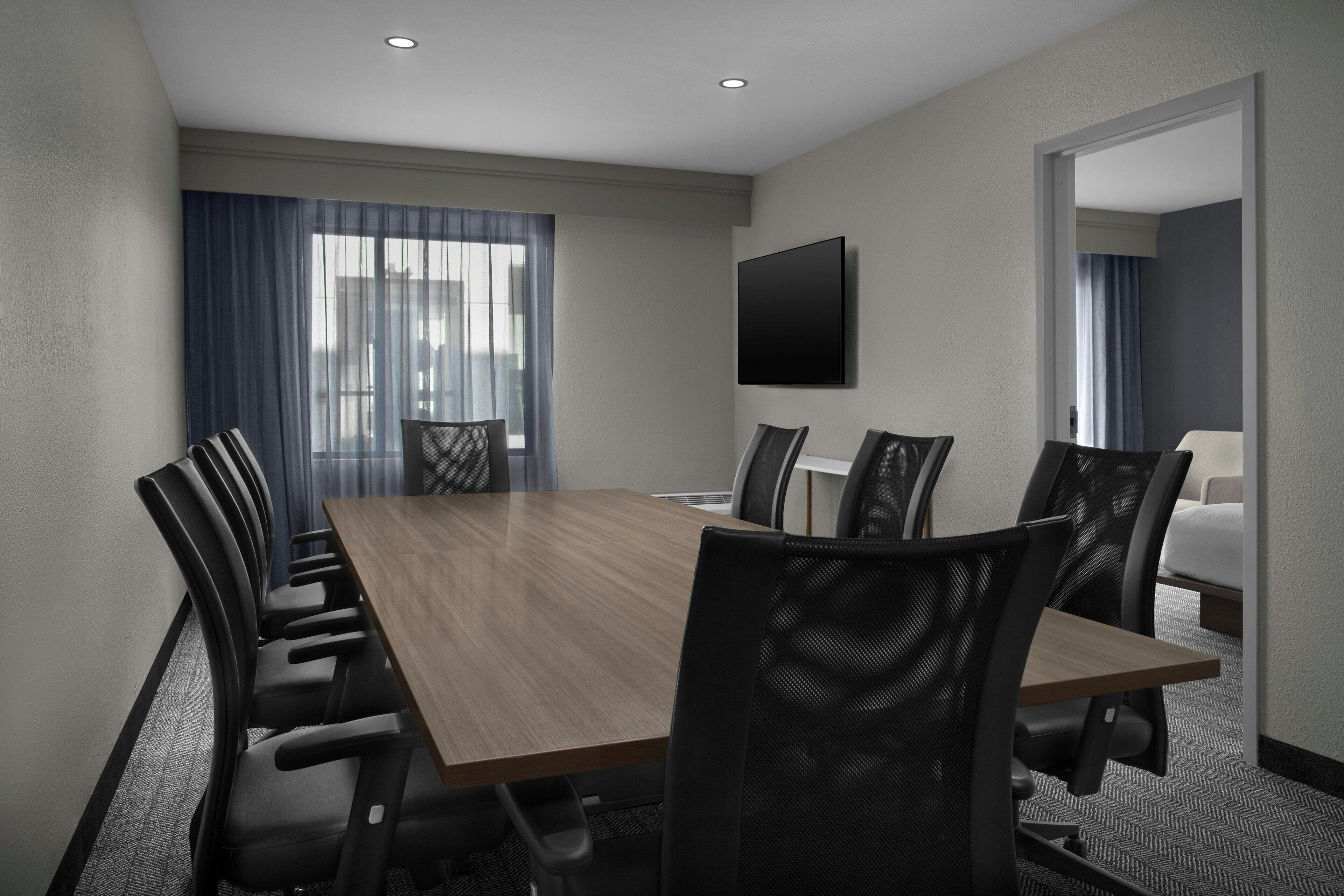 A conference room with a long table, six chairs, a flat-screen TV, and a large window with curtains.