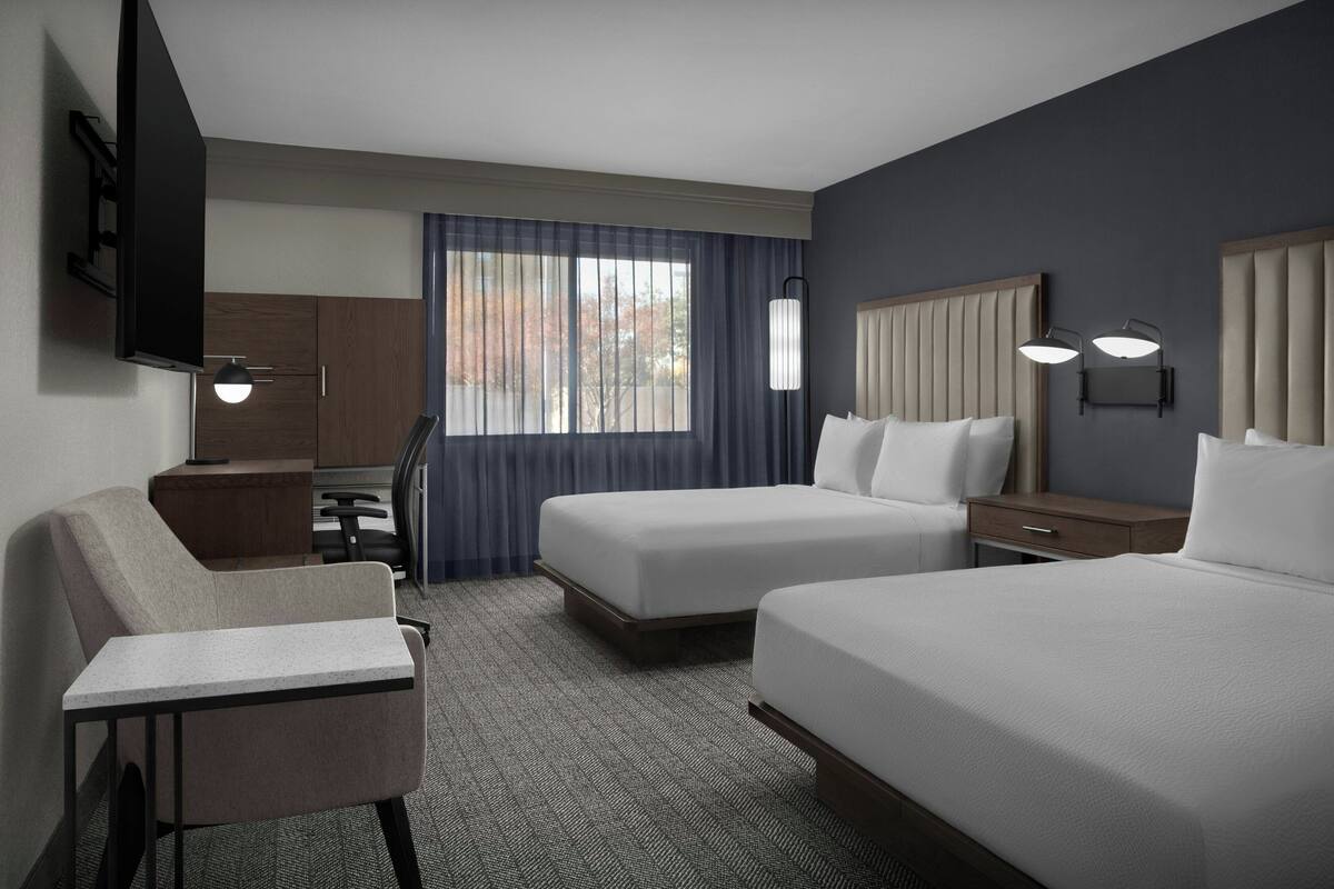 A hotel room with two beds, a desk, a chair, a TV, and a window with curtains.