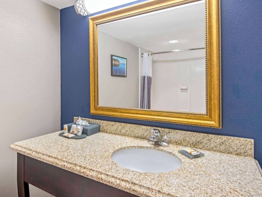 A bathroom with a granite countertop sink, a gold-framed mirror, and a small towel dispenser.