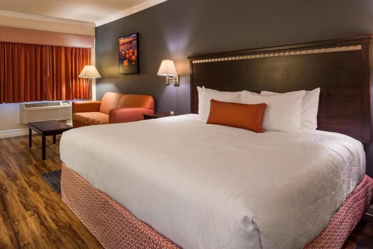 Quarto de hotel com uma cama grande, duas cadeiras, um abajur fixado na parede e um quadro.