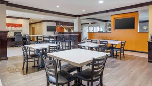 A spacious dining area with multiple tables and chairs, a kitchen in the background, and a large screen on the wall.