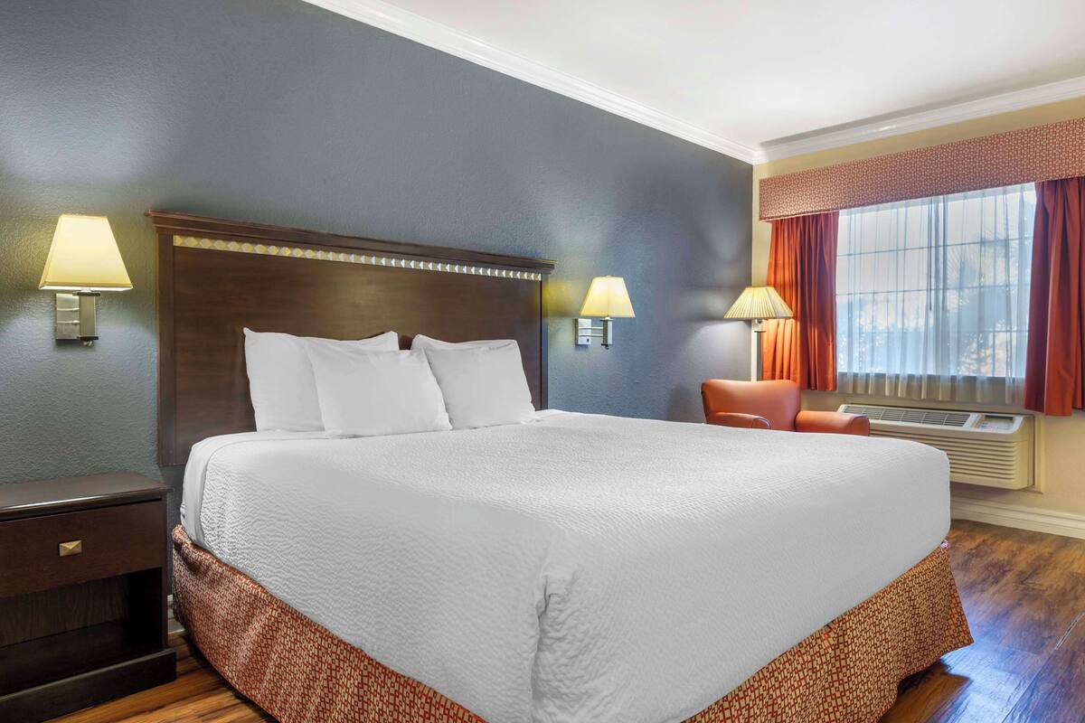Quarto de hotel com uma cama grande, duas luminárias de cabeceira, uma cadeira e uma janela com cortinas.