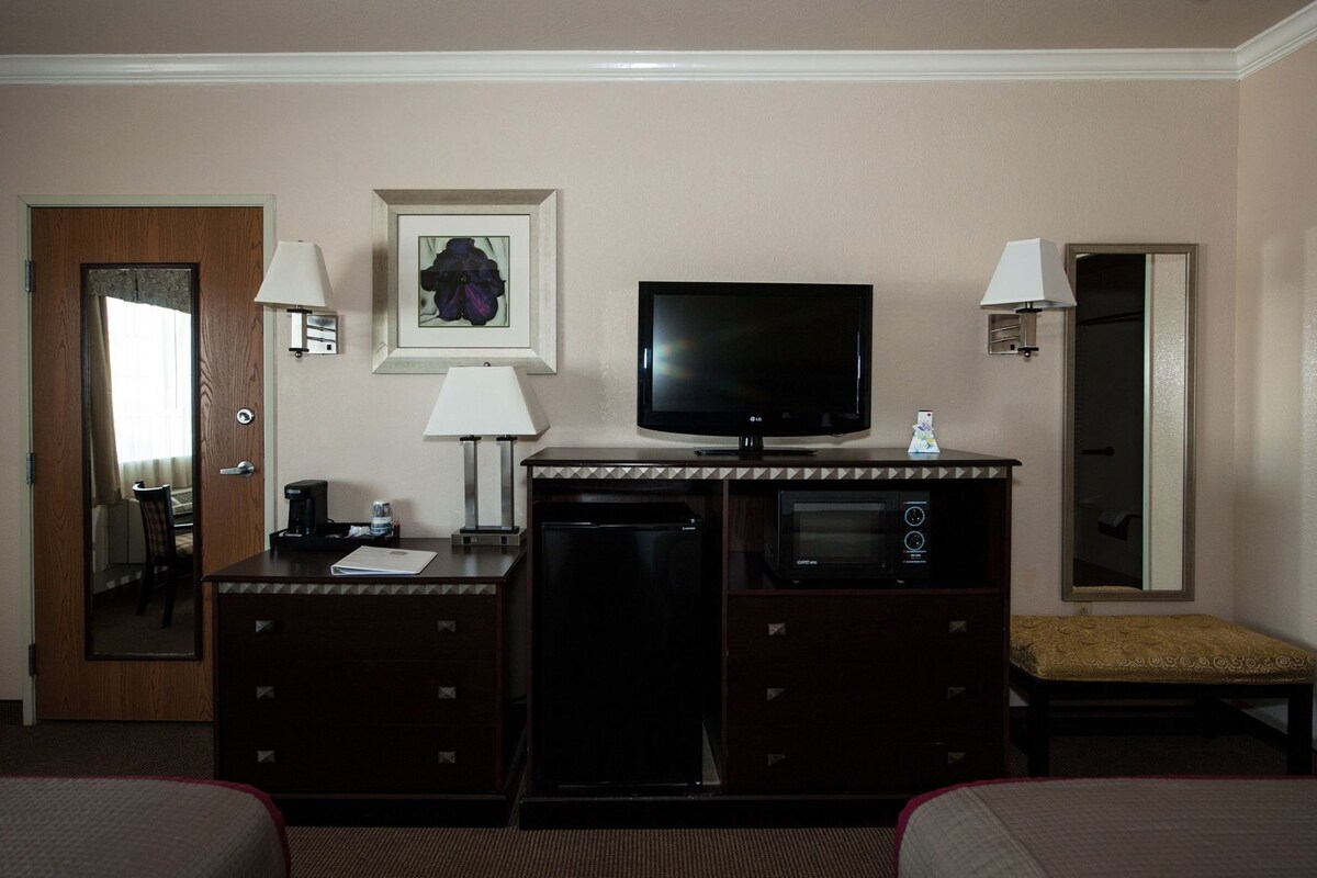 A hotel room with a TV, a desk with a lamp, a microwave, a small refrigerator, a bedside table, and a chair.