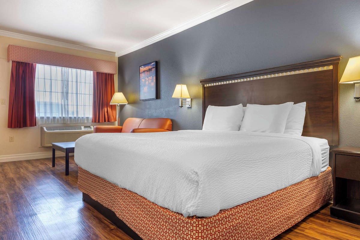 Quarto de hotel com uma cama grande, duas luminárias de cabeceira, uma mesa de cabeceira, uma cadeira e uma janela com cortinas.
