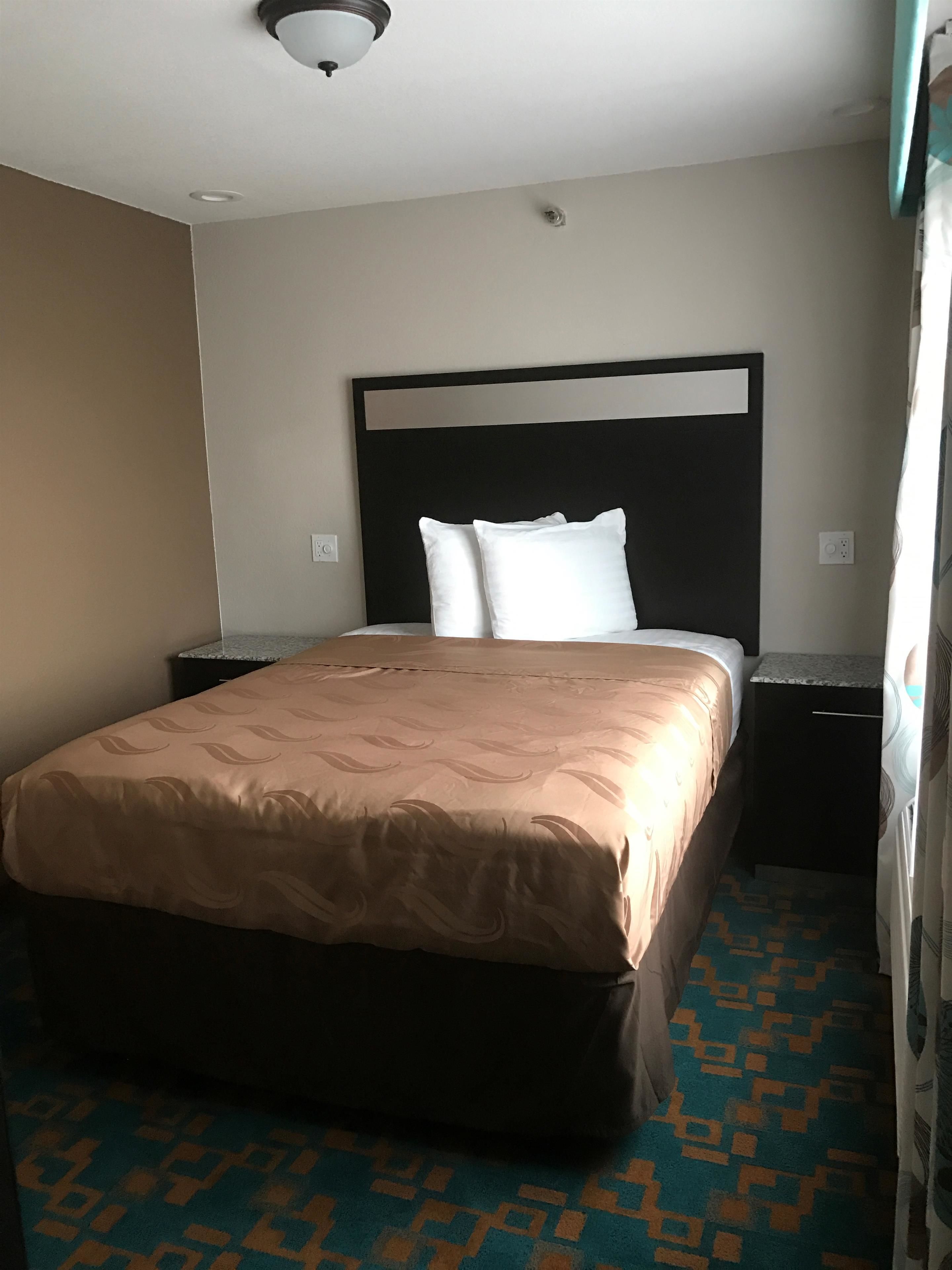 A hotel room with a bed, a headboard, a nightstand, and a patterned carpet.