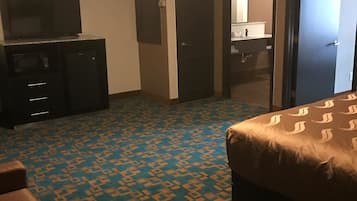 Habitación de hotel con alfombra estampada, una cama, un escritorio con televisor y un baño visible a través de una puerta abierta.