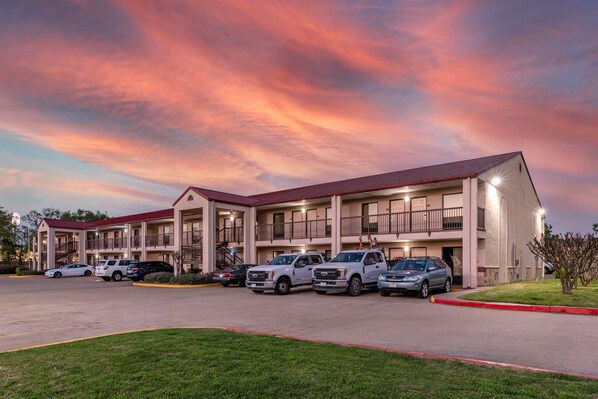 A multi-story hotel with a red roof, multiple balconies, and a parking lot with several SUVs.