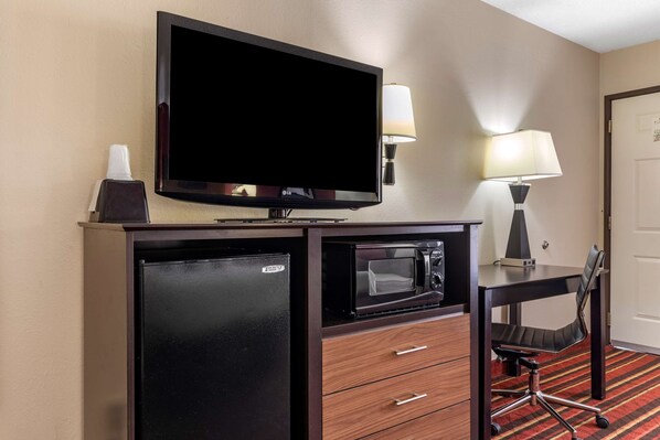 A compact hotel room with a flat-screen TV, a mini-fridge, a microwave, a desk with a chair, and a lamp.