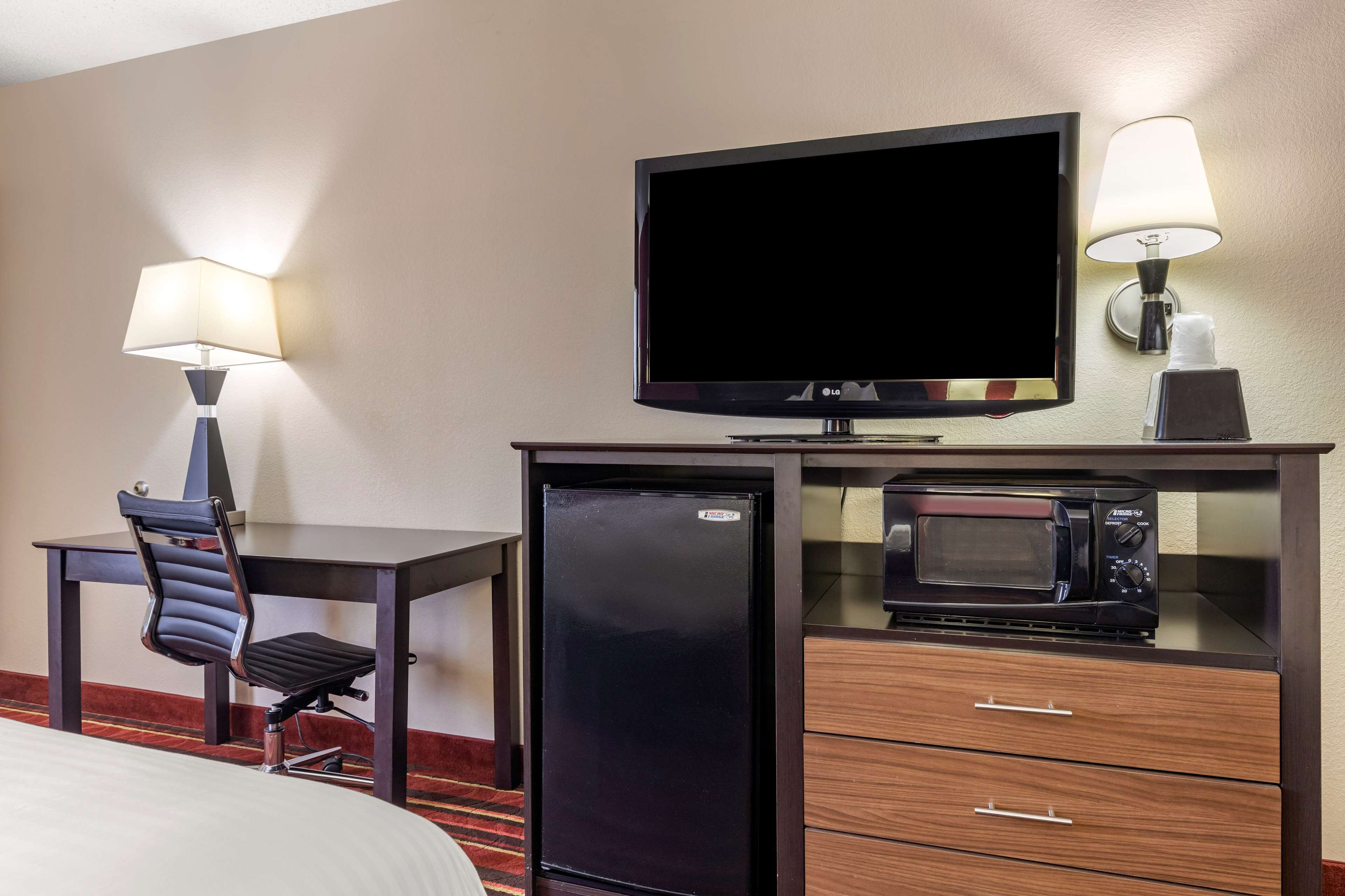 A hotel room with a bed, a desk with a chair, a television, a refrigerator, a microwave, and a lamp.