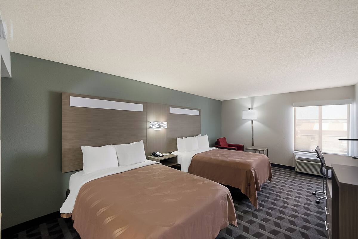 A hotel room with two beds, a desk, a chair, a lamp, and a window.