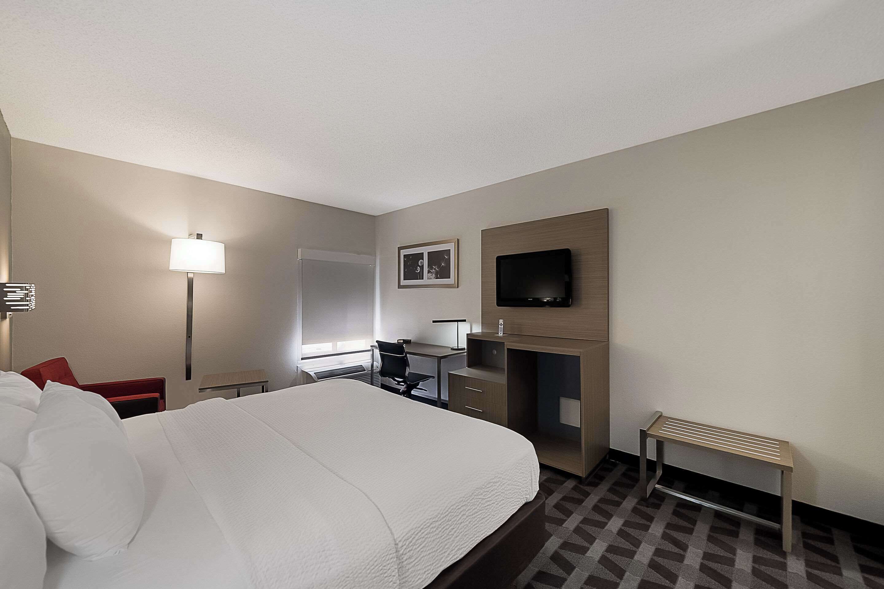 A hotel room with a large bed, a desk with a chair, a television mounted on the wall, and a small bench.